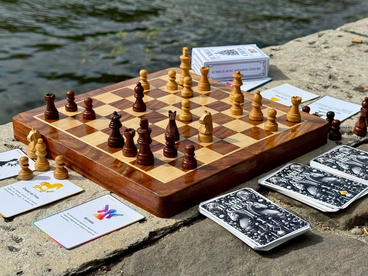 Échiquier en bois au bord de l’eau, entouré de cartes modificateurs et de la boîte Jumble Chess