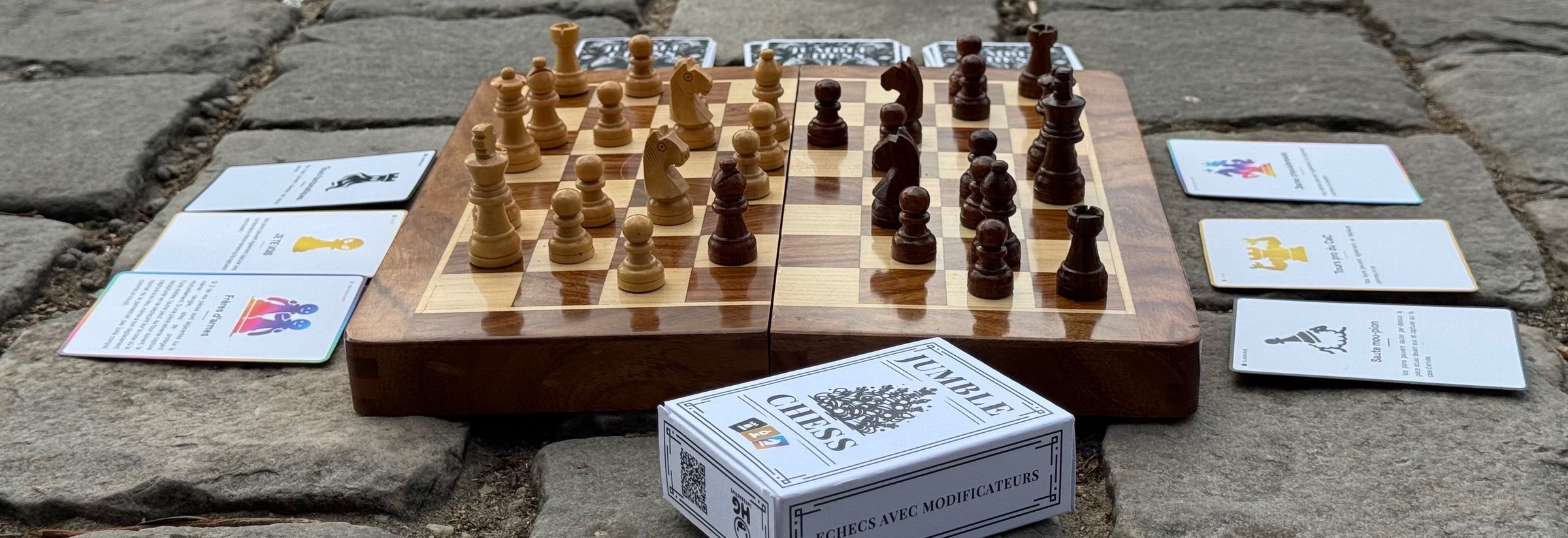 Échiquier en bois, cartes modificateurs et boîte Jumble Chess posés sur des pavés